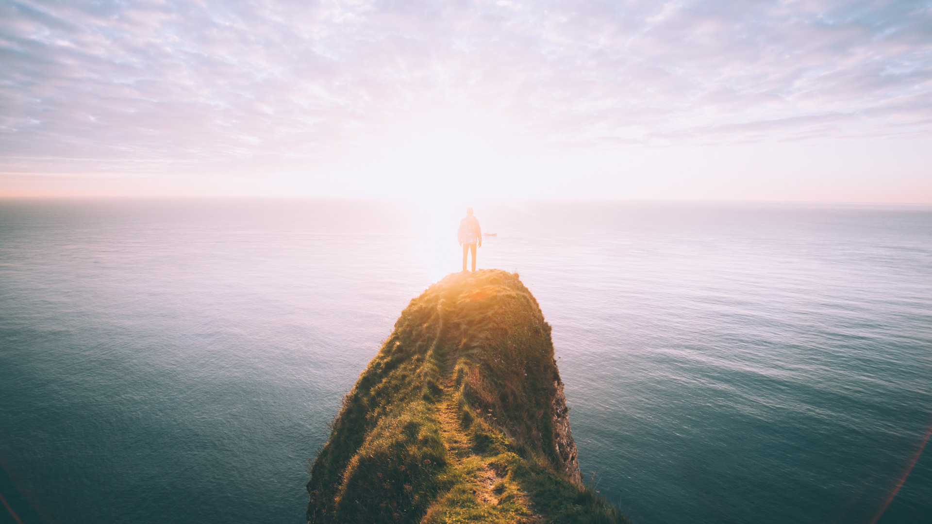 Licht am Horizont: Person steht auf einem Felsen über dem Meer, im Hintergrund geht die Sonne über dem Meer auf